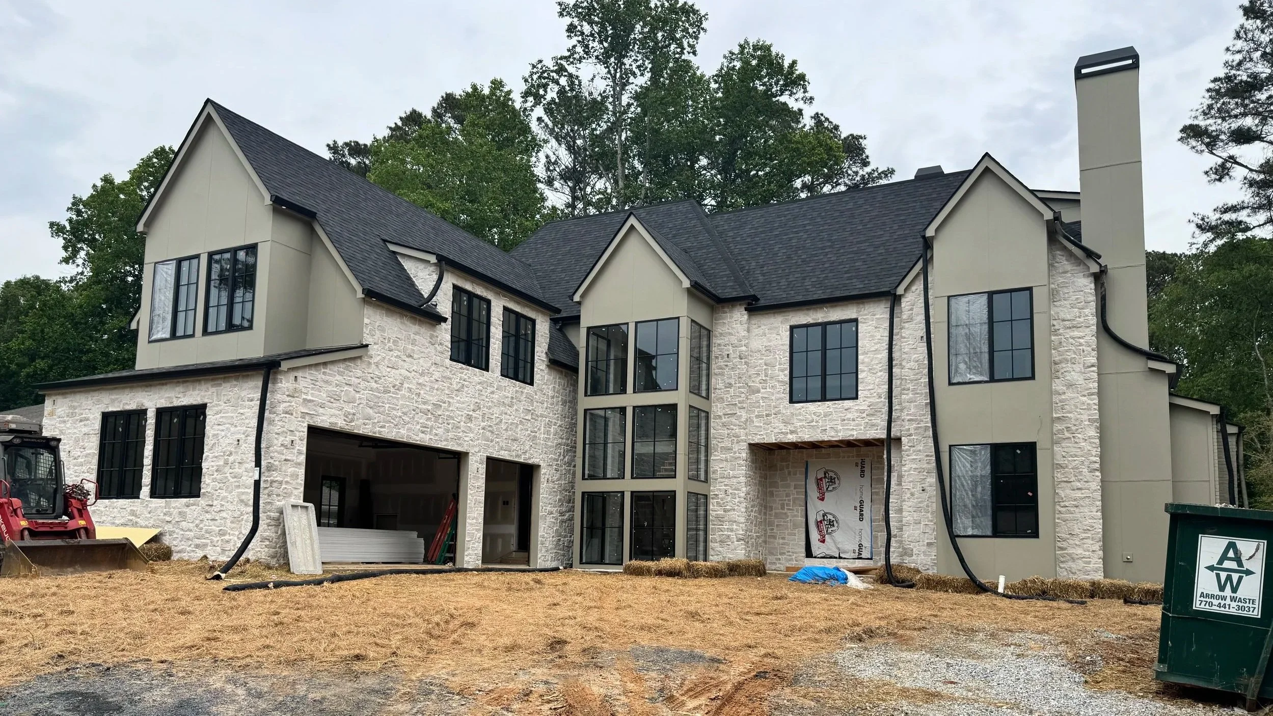 A large modern house under construction with white stone and beige siding exterior, multiple large windows, and a steep dark roof. Construction equipment and materials are in the foreground.