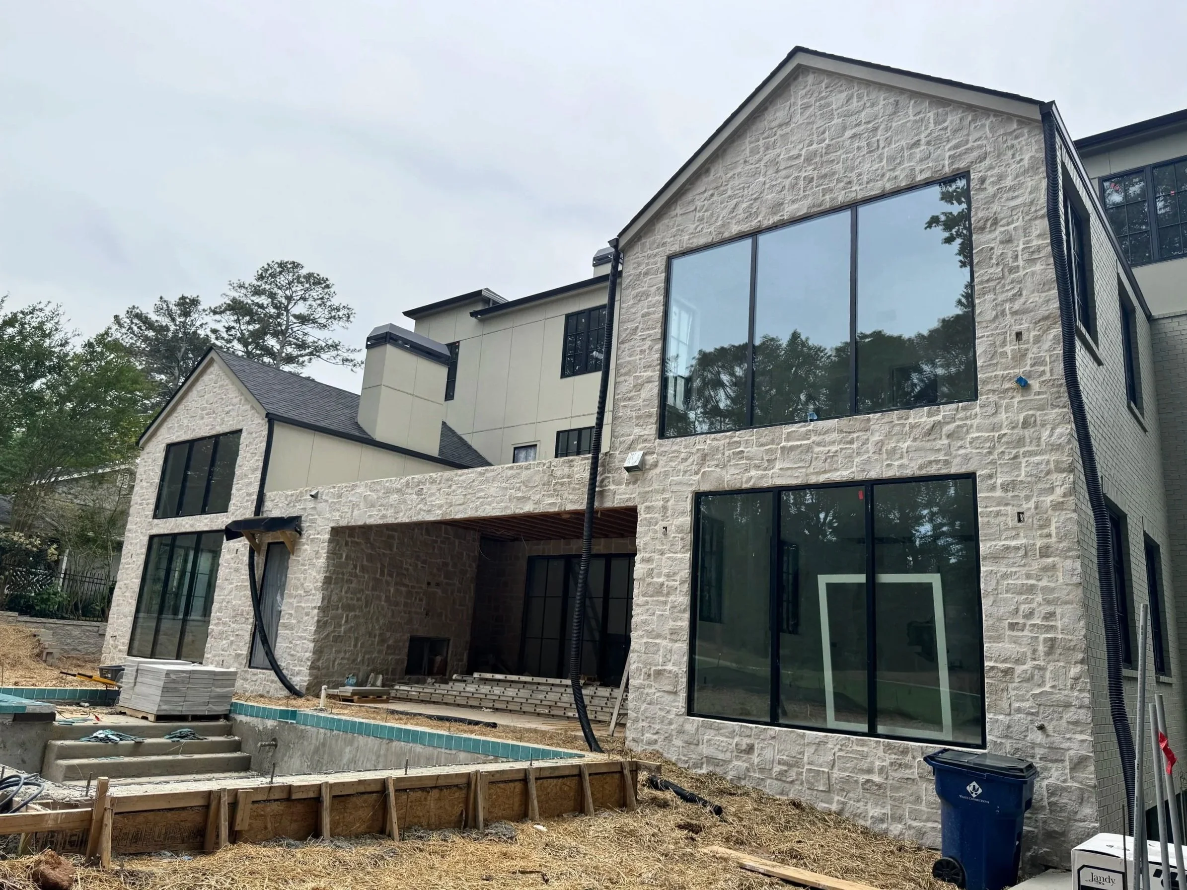 A multi-story modern house under construction with large glass windows, stone and siding exterior, and a yard with construction material and tools.