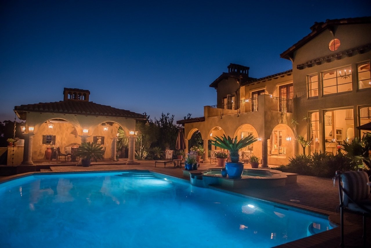 Luxury house with arched windows and balconies illuminated at night, swimming pool in the foreground, and potted plants around the pool area.