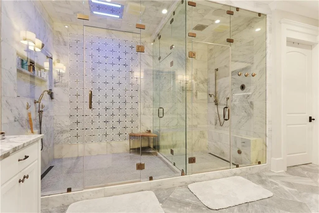 Elegant bathroom with a large glass-enclosed shower featuring marble walls, a built-in bench, and a decorative tile wall. There's a white vanity with a marble countertop and two white rugs on the grey marble tile floor.