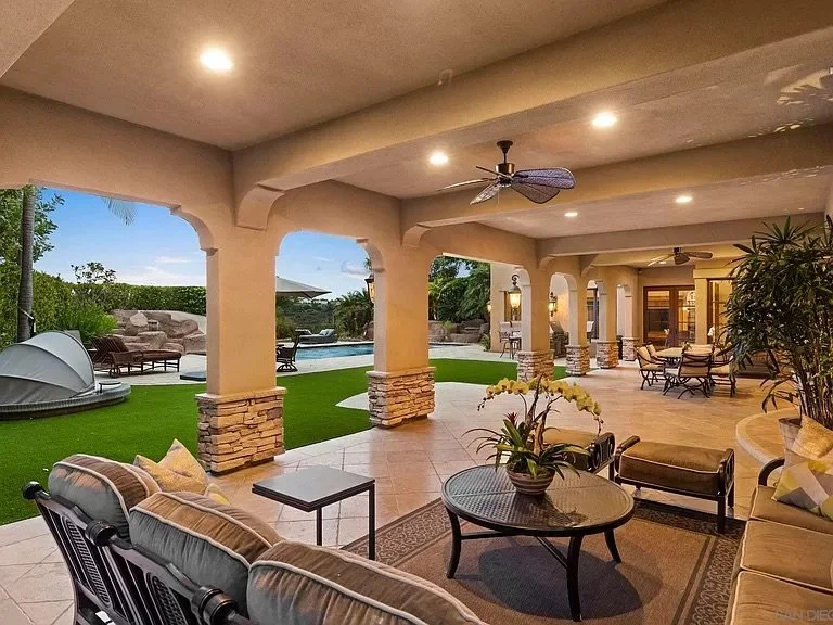 Covered outdoor patio with beige columns, ceiling fans, and outdoor furniture overlooking a backyard with a pool, grass, trees, and lounge chairs.