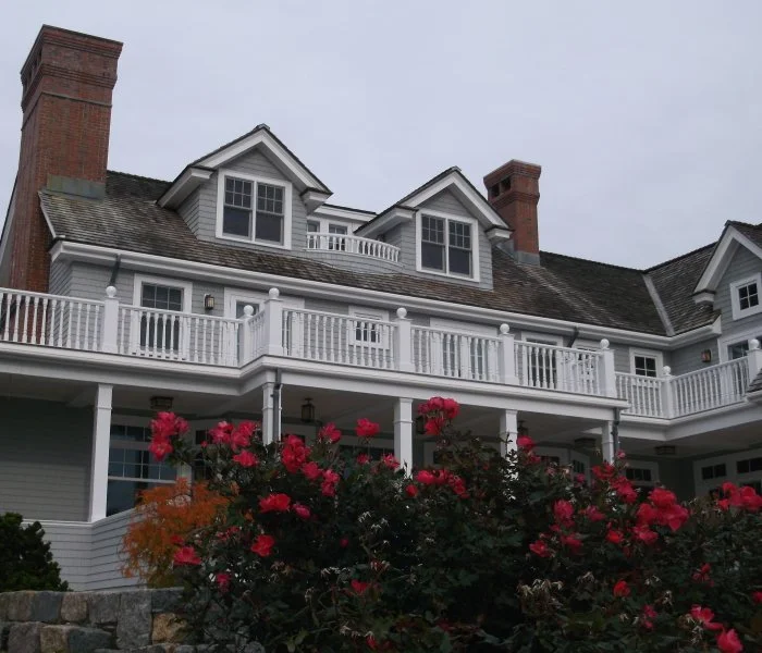 Large white house with multiple gabled windows, a wrap-around balcony, and brick chimneys. Pink flowers and greenery in the foreground.