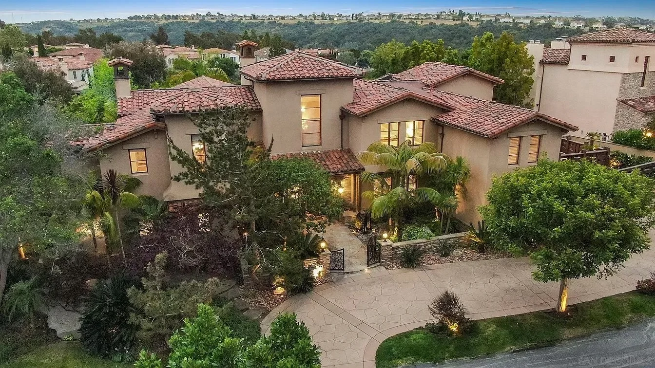 Aerial view of a large house with a tiled roof, surrounded by trees and greenery, with a spacious driveway and landscape lighting at dusk.
