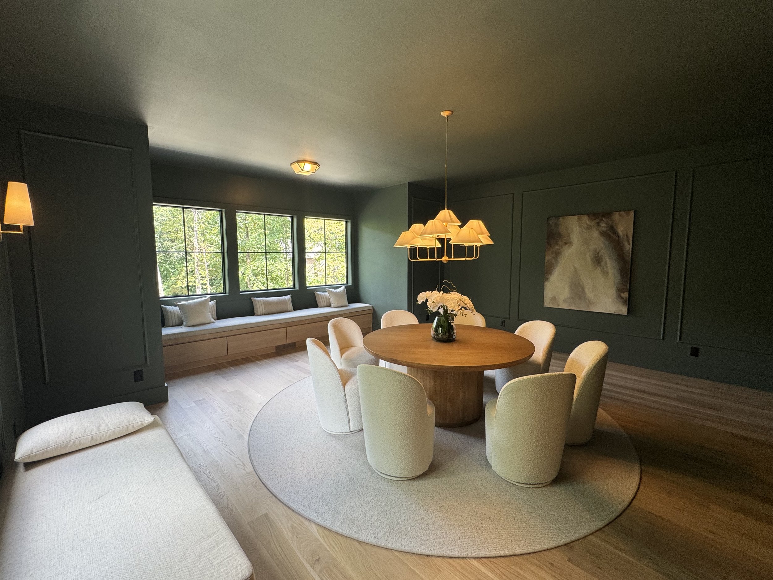 A dining room with a round wooden table and cream-colored upholstered chairs, a built-in window seat with white pillows, dark gray walls with decorative molding, large windows with greenery outside, and modern light fixtures, including a chandelier a