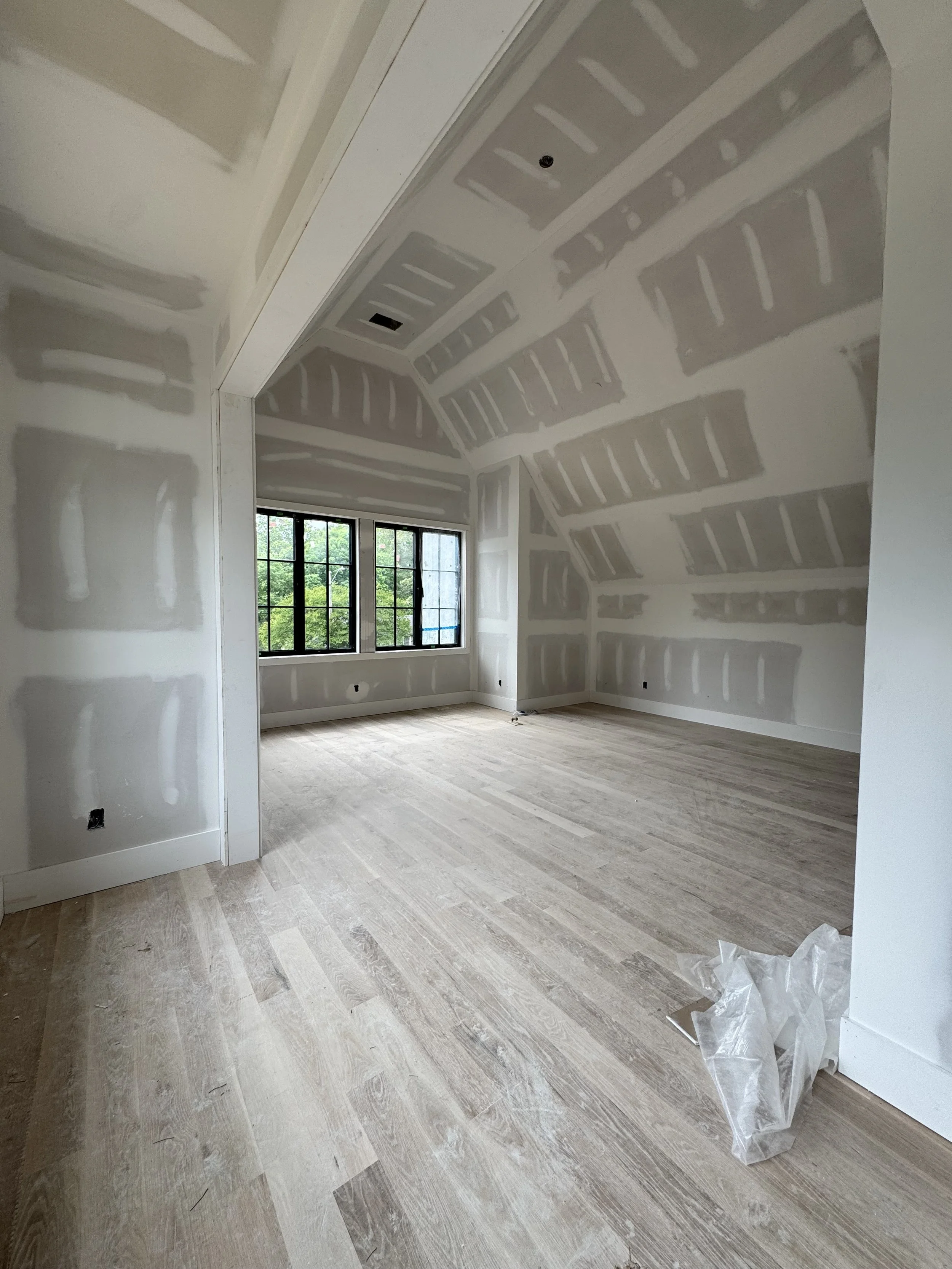 A room under construction with drywall installed and unpainted. The ceiling has drywall with visible joint seams and a central recessed light opening. The floor is unfinished with wood-looking planks, and there is a large window showing greenery outs