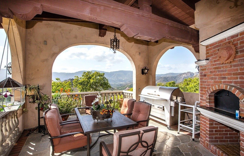 Outdoor covered patio with a view of mountains, featuring a dining table with six chairs, a BBQ grill, brick and stucco walls, arches, and decorative lanterns