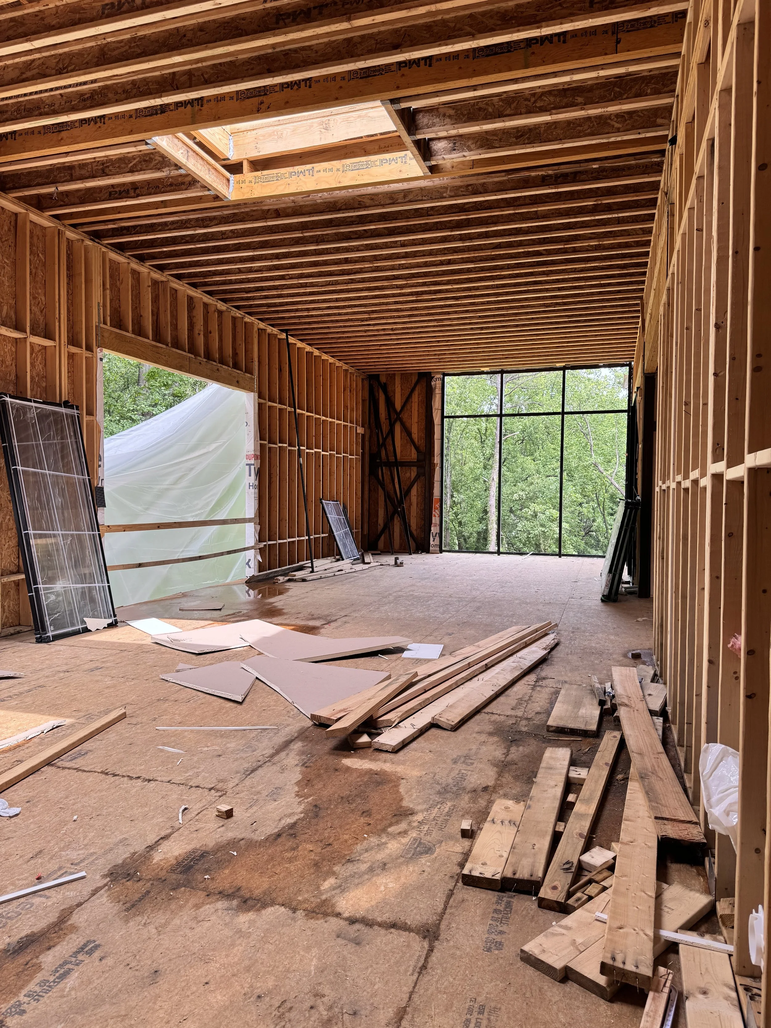 Interior view of a house under construction with exposed wooden framing, some materials and tools scattered on the floor, and large windows showing green trees outside.