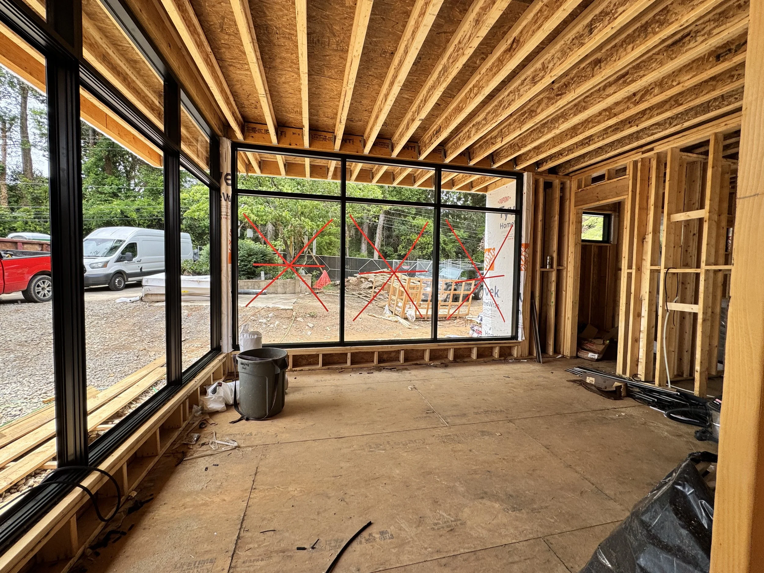 Interior view of a house under construction showing wooden framing, large window openings, and construction materials inside and outside.