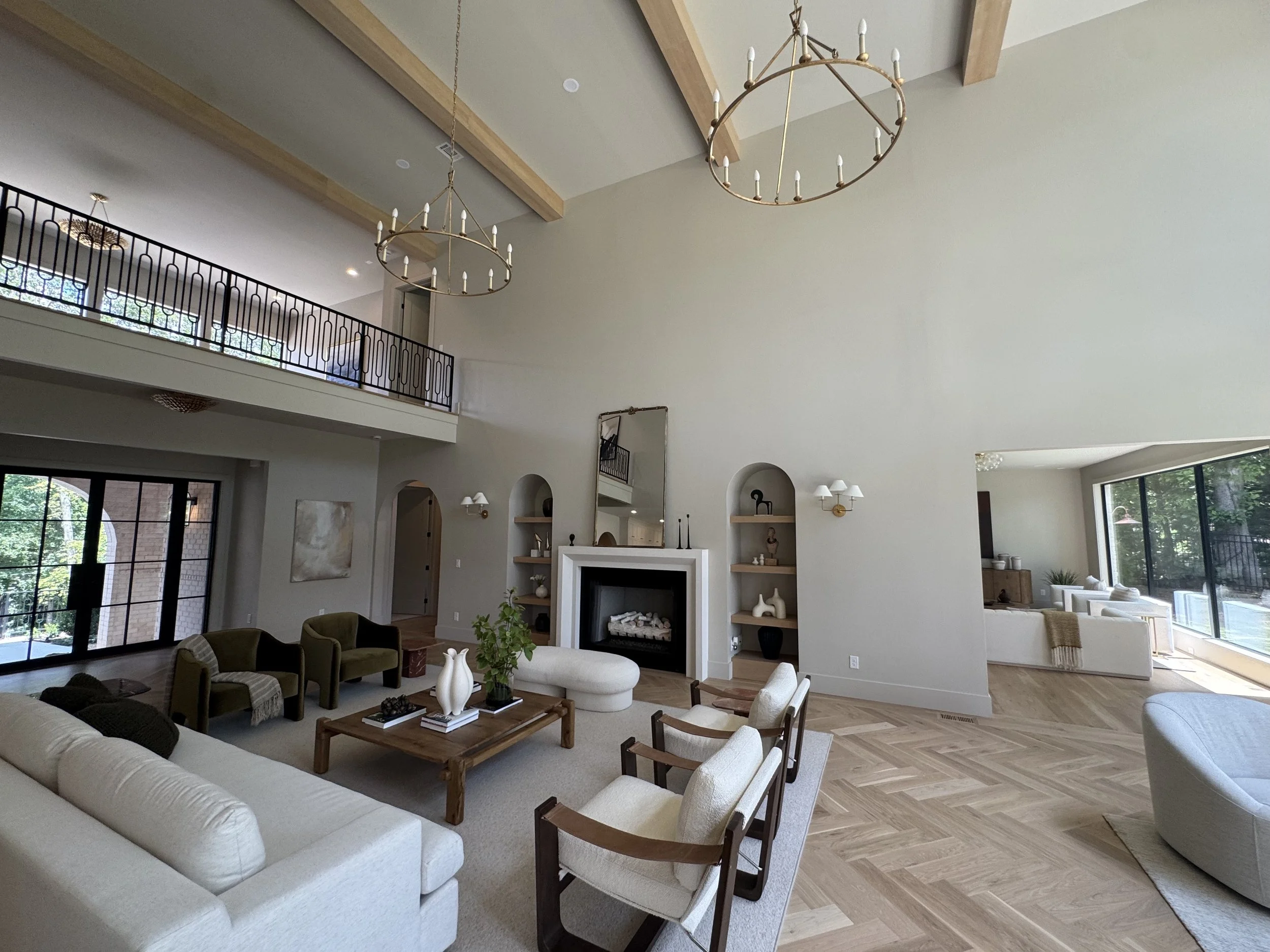 Spacious living room with white walls, large windows, modern furniture including sofas, chairs, and a coffee table, a fireplace with decorative items, and high ceilings with wooden beams and chandelier lighting.