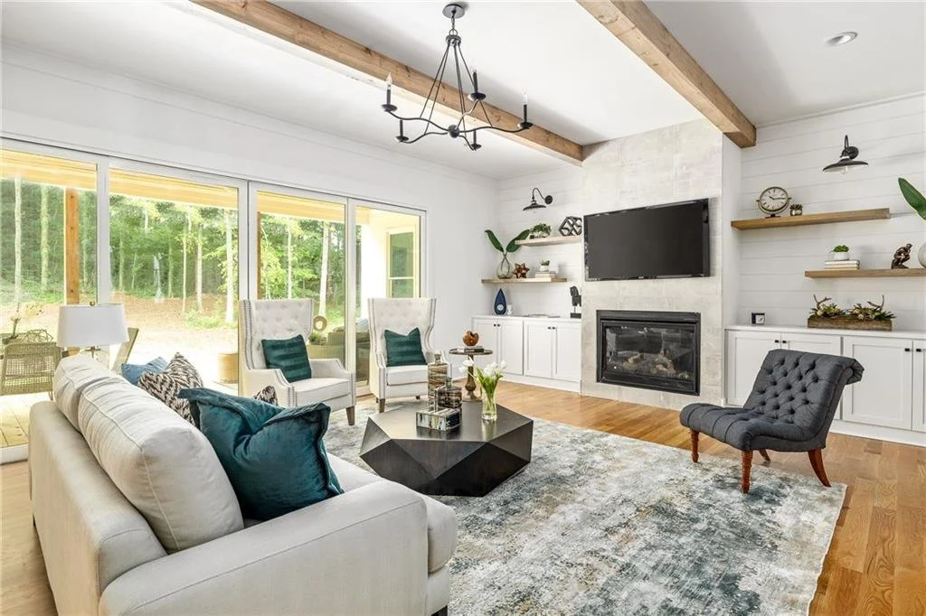 Bright living room with large sliding glass doors showing a wooded yard outside. Features a white sofa with blue and patterned pillows, two white armchairs with dark teal pillows, a gray tufted accent chair, a dark geometric coffee table, a stone fir