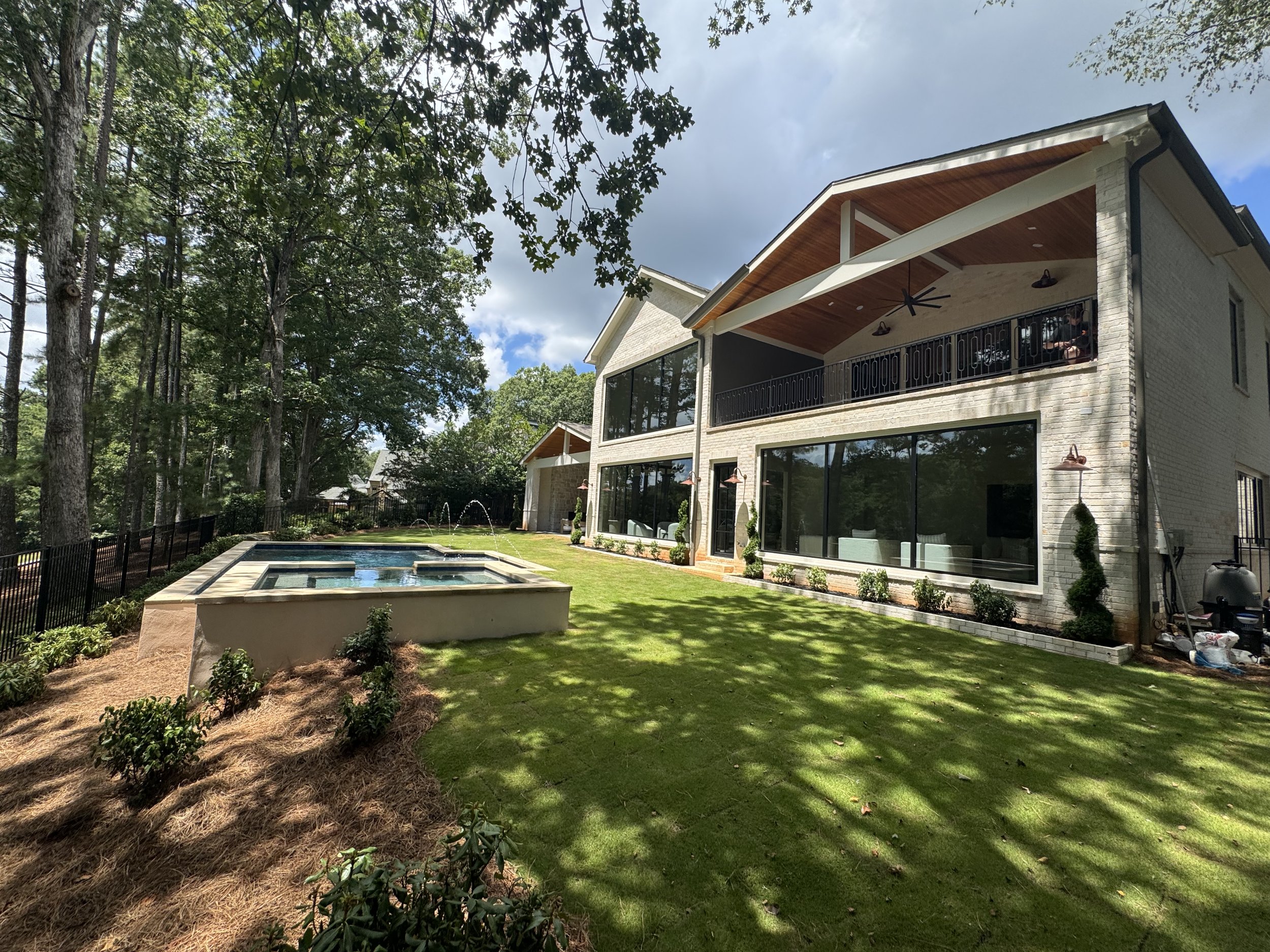 View of a backyard with a modern house featuring large windows, a balcony, and a wooden ceiling. The yard has a swimming pool, surrounding landscaping, and tall trees providing shade, with a partly cloudy sky overhead.
