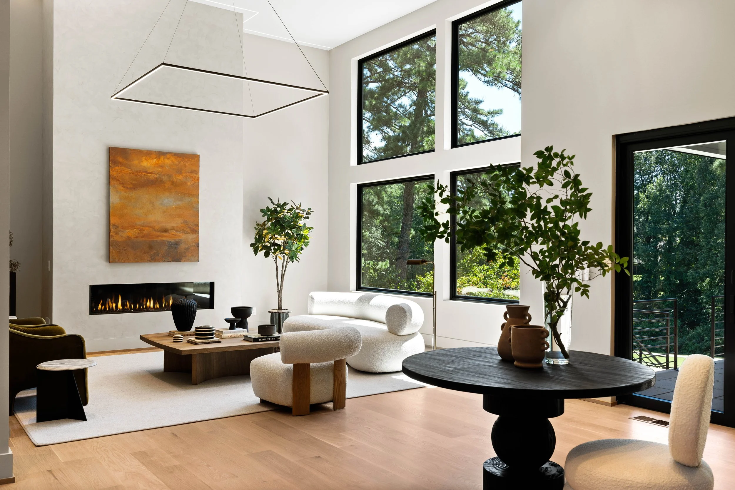 Modern living room with large windows showing trees outside, white walls, a built-in fireplace, and various contemporary furniture and plants.