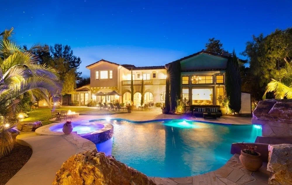 Night view of a luxurious backyard with a swimming pool illuminated by underwater lights, surrounded by palm trees and a large house with lit windows.