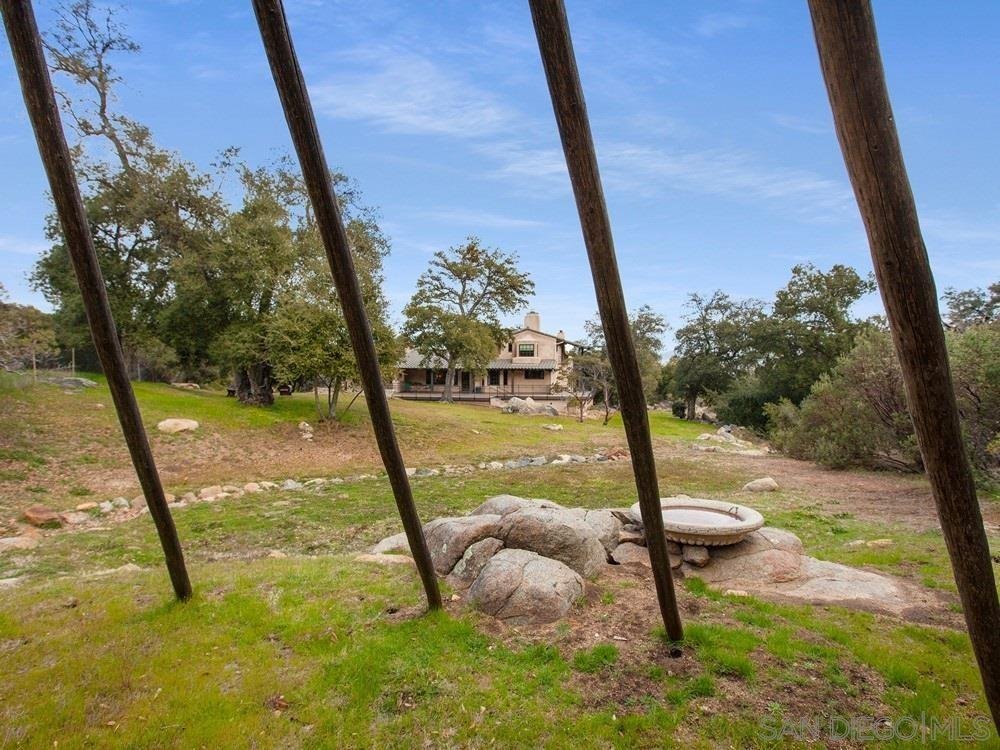 View of a house in the distance through wooden poles, with a grassy yard, rocks, trees, and an empty stone birdbath in the foreground.