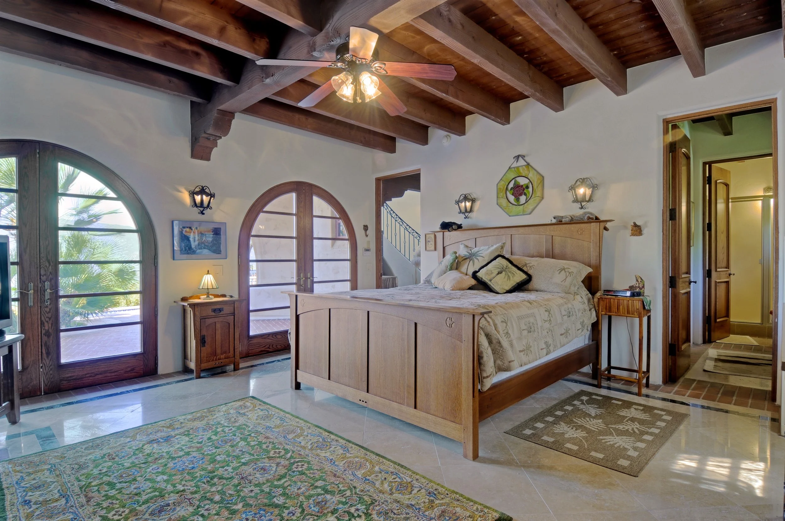 A bedroom with a wooden bed, tropical-themed pillows, a nightstand with a lamp, and two wall-mounted lanterns. The room has wooden-beamed vaulted ceiling, arched glass doors leading outside, and a colorful area rug.