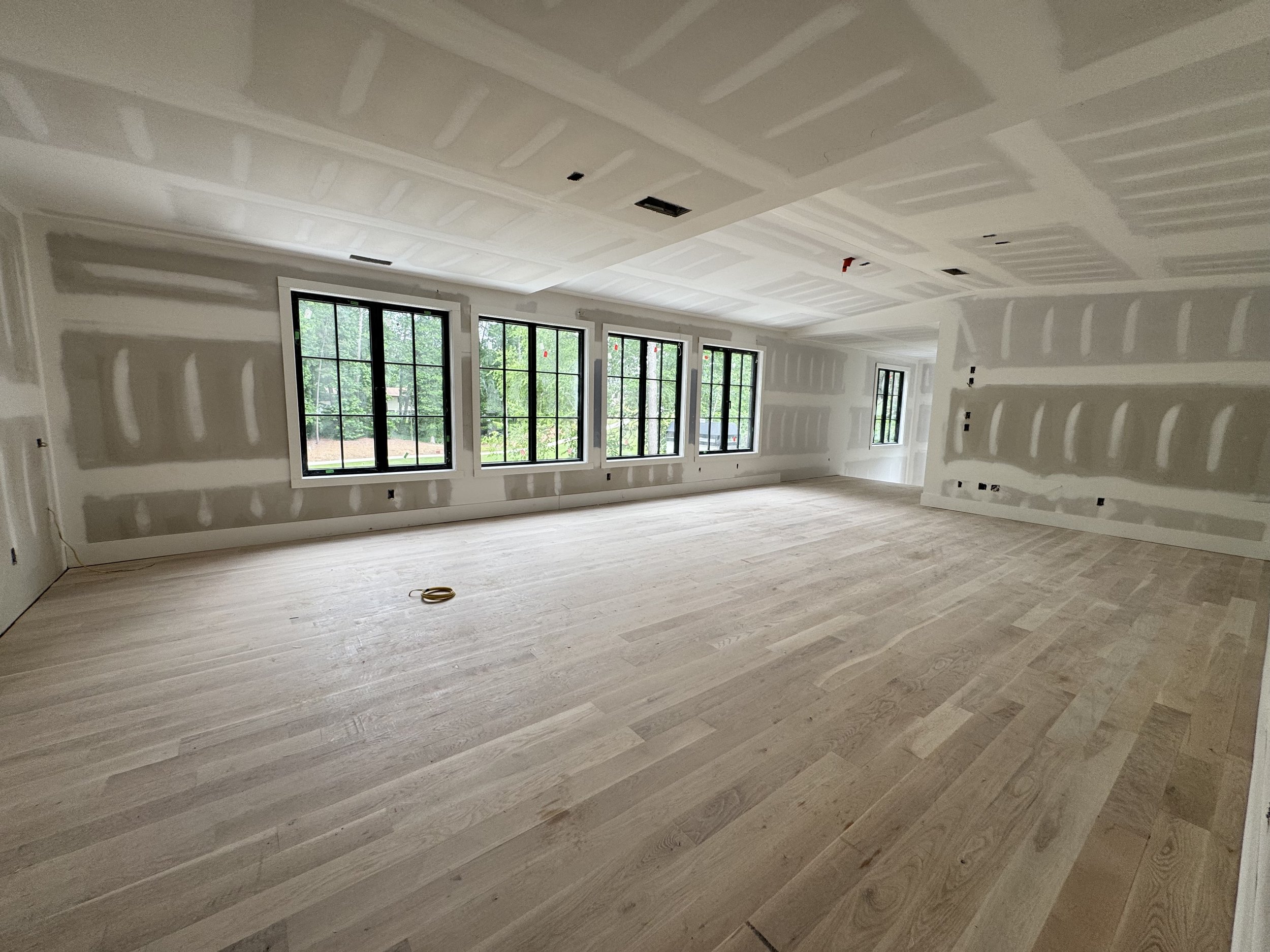 Room under construction with drywall joints taped and ready for painting, large windows showing greenery outside, and wooden flooring.