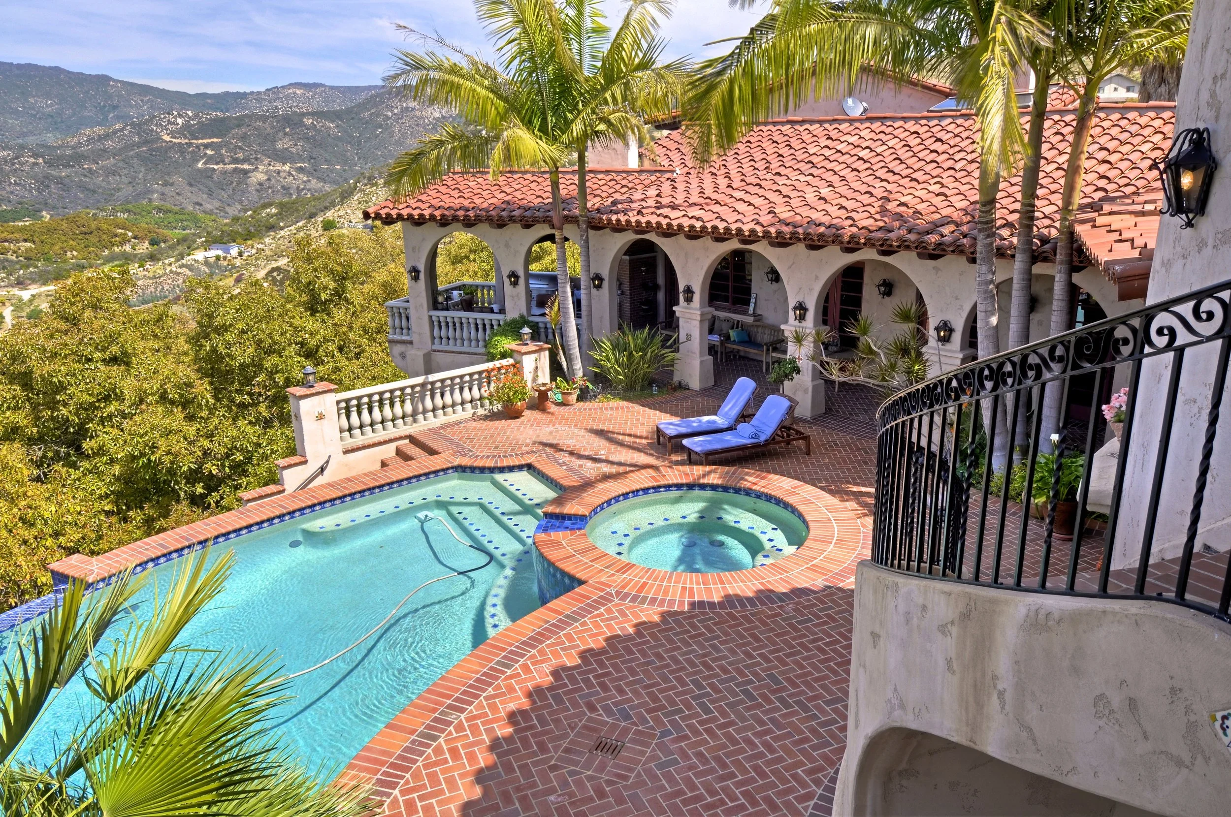 A house with a red tile roof, surrounded by palm trees, overlooking a mountainous landscape. The backyard features a swimming pool and hot tub with red brick paving, outdoor seating, and a black wrought iron fence.