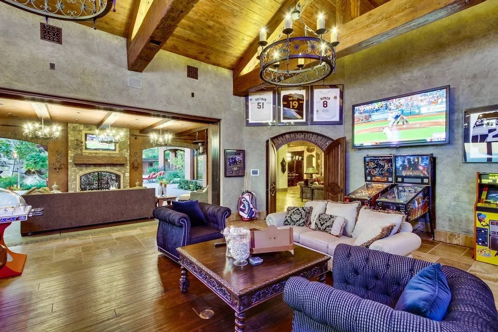 Living room with wooden ceiling beams, large TV showing a baseball game, arcade game machines, a white sofa with patterned pillows, dark armchairs, a wooden coffee table, and sports jerseys framed on the wall.