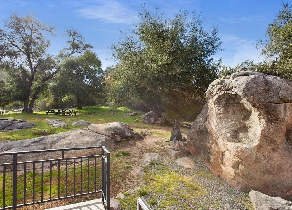 A natural park scene with large rocks, green grass, trees, a picnic table, and a railing in the foreground, under a blue sky with light clouds.