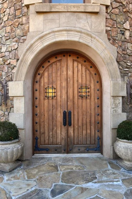 Wooden double door with iron hardware, set within an arched stone doorway, flanked by large stone planters with green bushes.