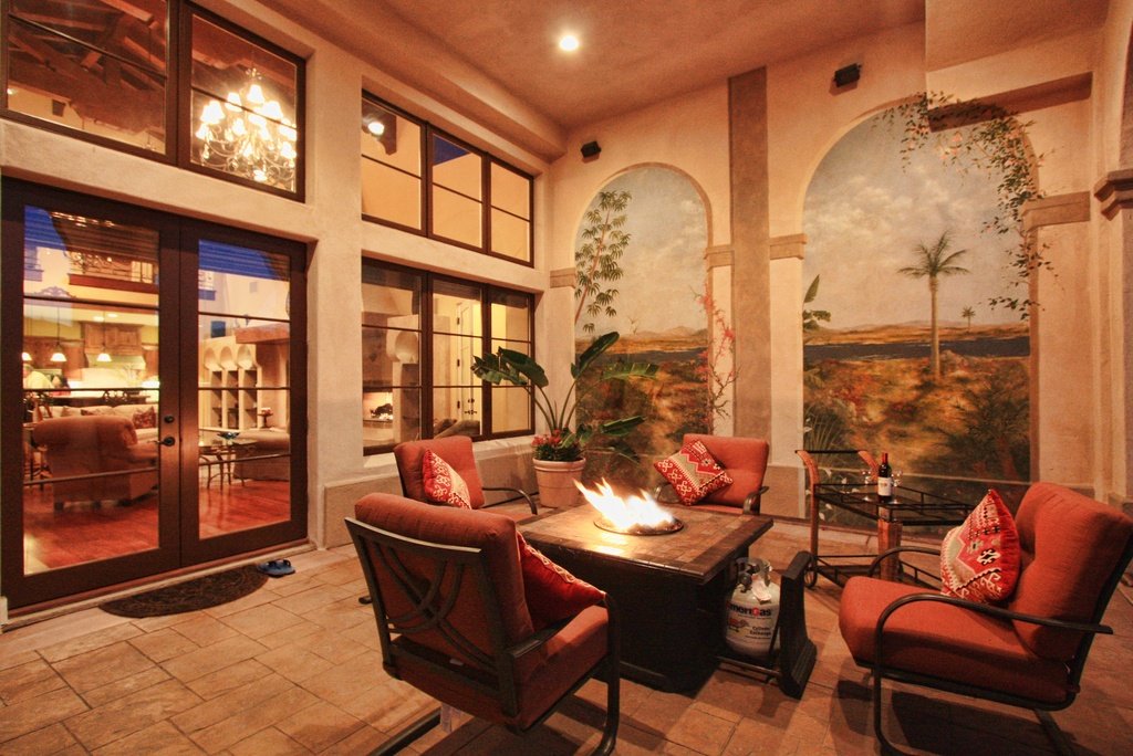 A cozy outdoor patio area with four cushioned chairs around a fire pit, decorated with red pillows, and potted plants. The background features large windows and a mural of a desert landscape with palm trees.