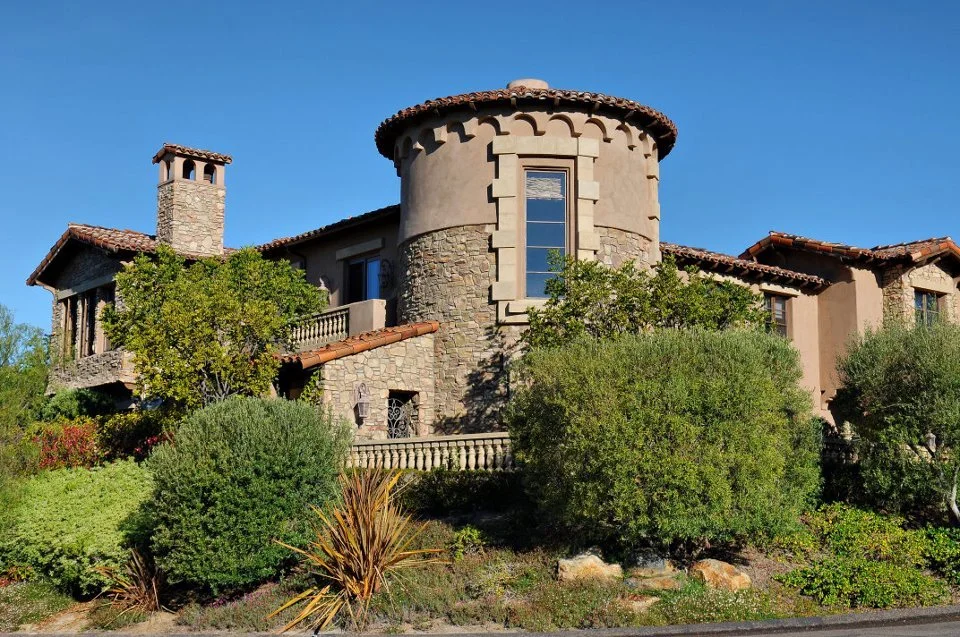 A large house with a stone and stucco facade, a round turret, a chimney, and surrounded by lush green bushes and trees under a clear blue sky.