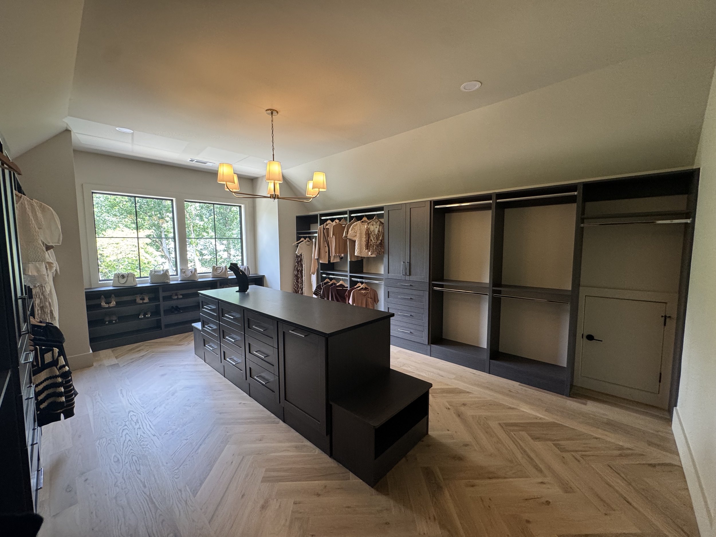 Inside a walk-in closet with a large central island, dark cabinetry for storage, and a window letting in natural light; clothes are hung on the right side, and handbags are placed on a shelf below the window.