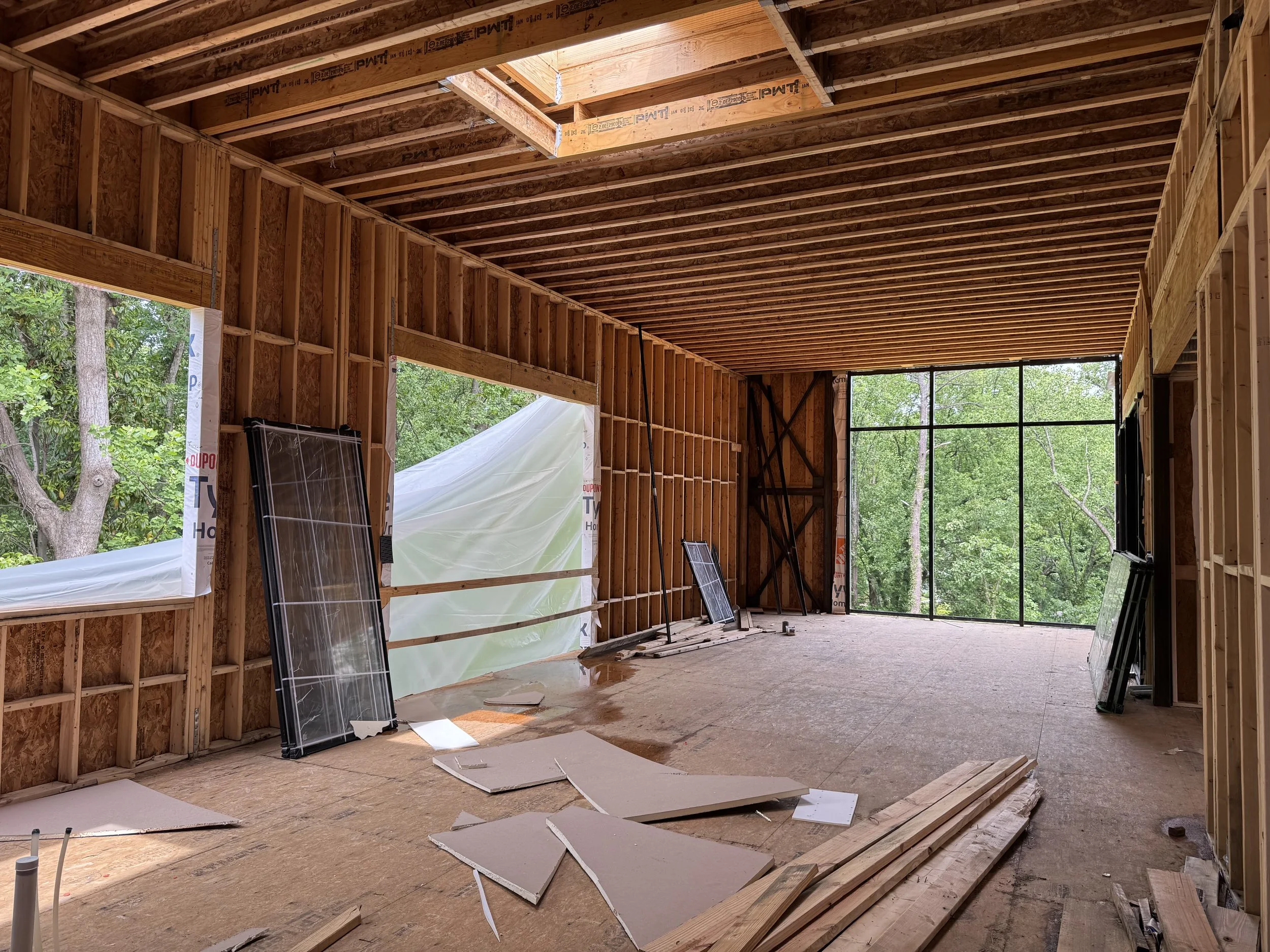 Interior view of a building under construction with exposed wooden framing, large open window, and construction materials scattered on the floor.