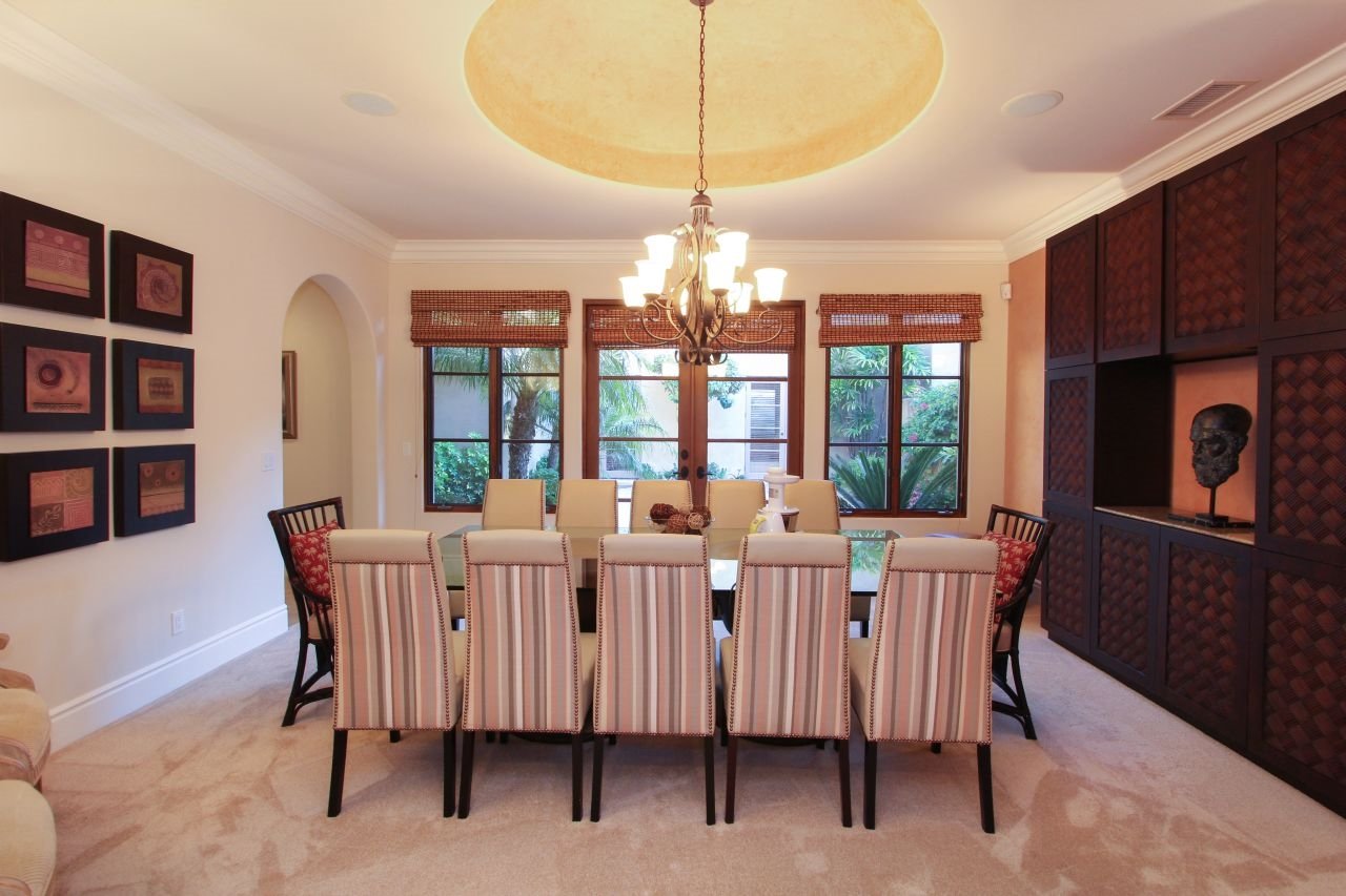A dining room with a long rectangular table surrounded by striped upholstered chairs. A chandelier hangs above the table. The room has windows with wooden blinds and views of greenery outside. There are framed artworks on one wall and a large dark wo