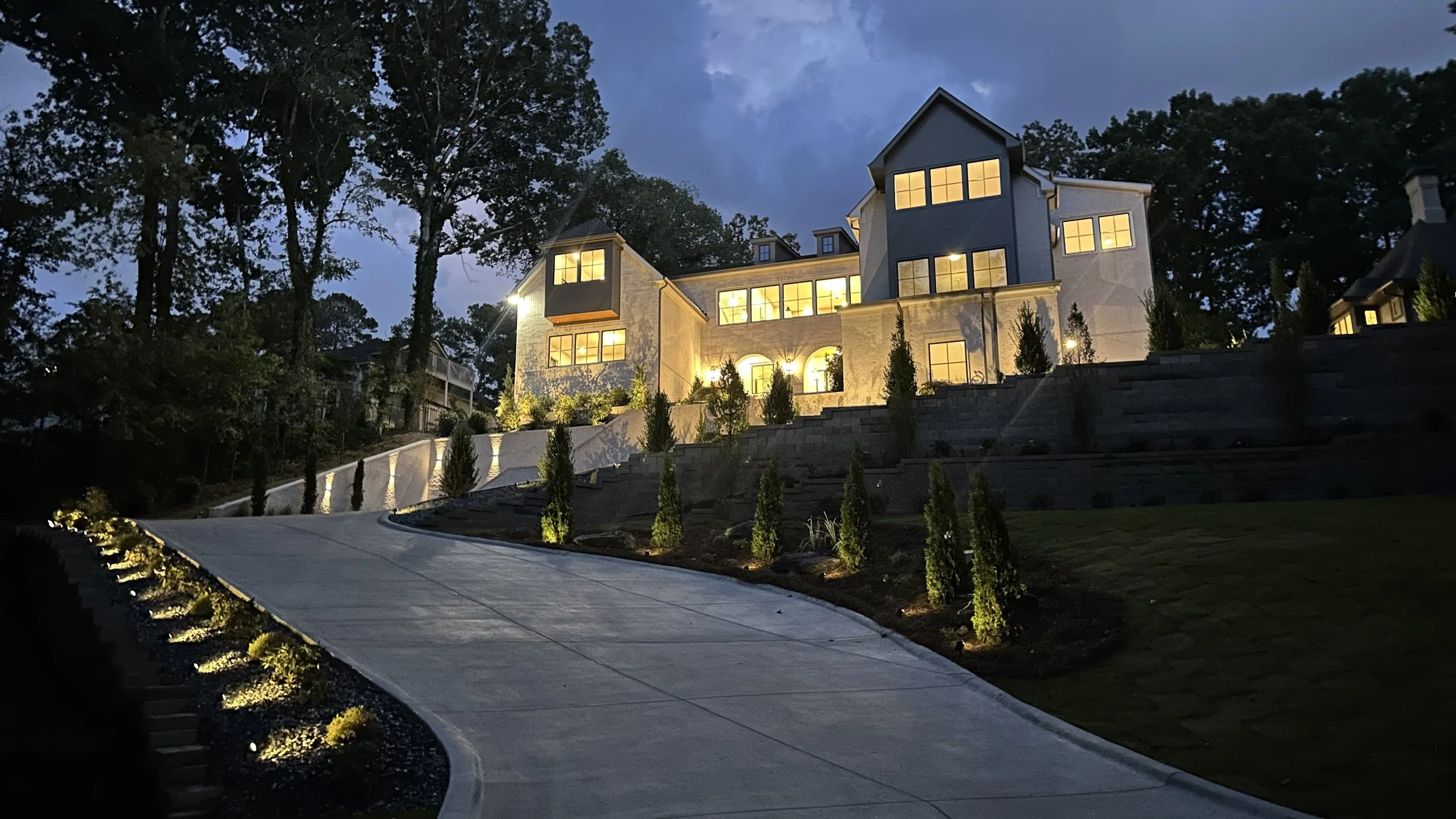 A multi-story house at dusk with lit windows, situated on a incline with landscaped yard, stone retaining walls, and a curved concrete driveway, surrounded by tall trees.