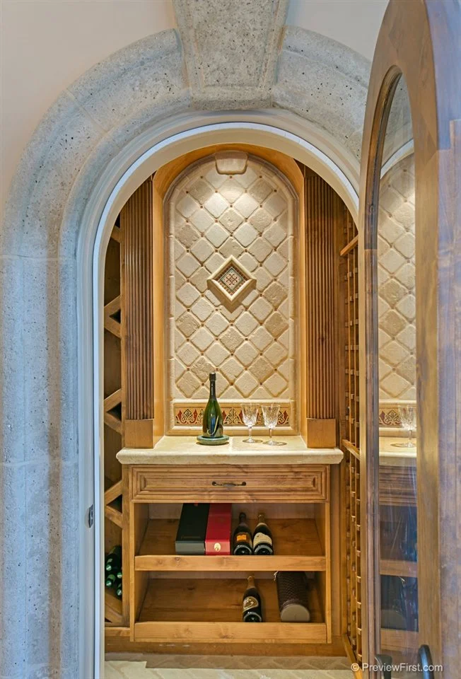 Wine storage area with wooden shelves, a glass bottle on a tray, two wine glasses, and wine bottles stored on lower shelves, framed by an arched stone doorway.