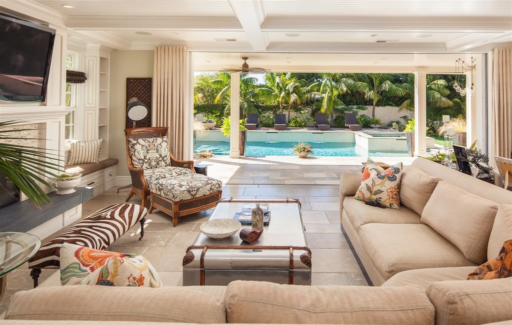 Living room with beige sofas, patterned armchair, and ottoman, overlooking a backyard with a swimming pool and lush green plants.