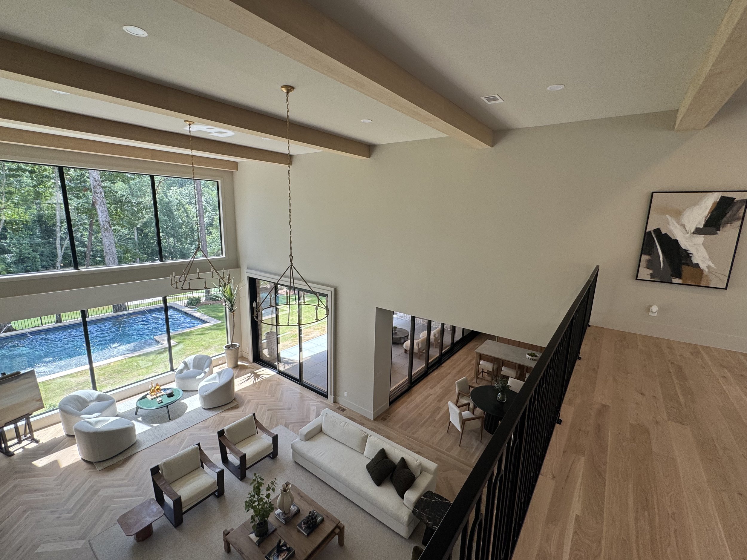 A spacious, modern living room with large windows overlooking a backyard with a swimming pool and tall trees. The room features light-colored walls, a high ceiling with wooden beams, a chandelier, and a mix of contemporary white and wooden furniture 