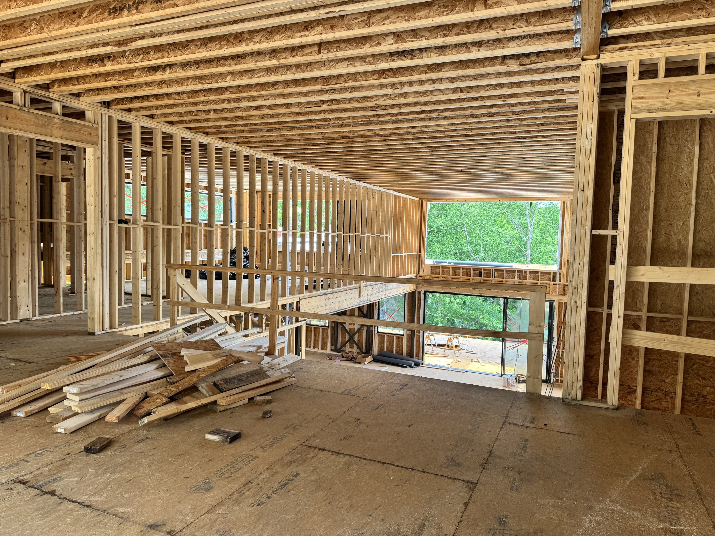 The interior of a house under construction with exposed wooden framing, floor, and ceiling, and a large window showing green trees outside.