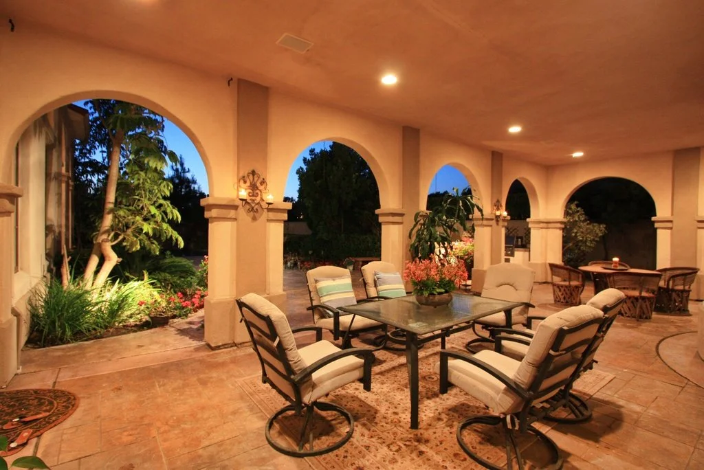 Covered patio area with outdoor furniture, including a table with a flower arrangement and cushioned chairs, illuminated by ceiling lights during evening time.