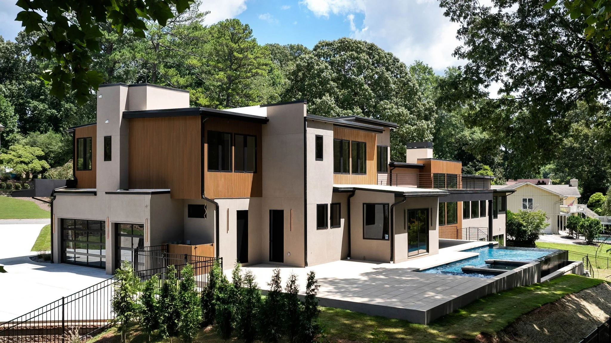 Modern multi-story house with large windows, wood paneling accents, a backyard with a swimming pool, and surrounded by green trees under a partly cloudy sky.