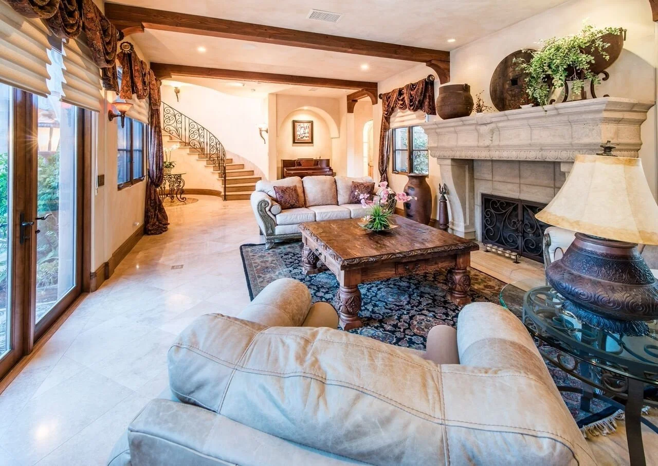 A spacious living room with a stone fireplace, a beige sofa, a wooden coffee table with a plant, and large windows with drapes, hardwood beams on the ceiling, and a staircase in the background.