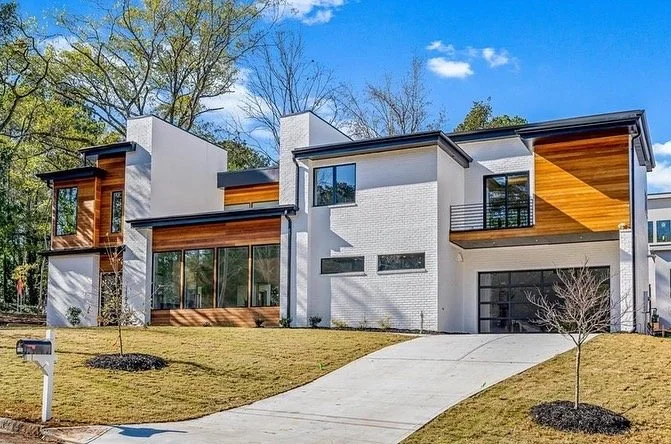 Modern multi-story house with white walls, wooden accents, large windows, and a sloped driveway in a landscaped yard.