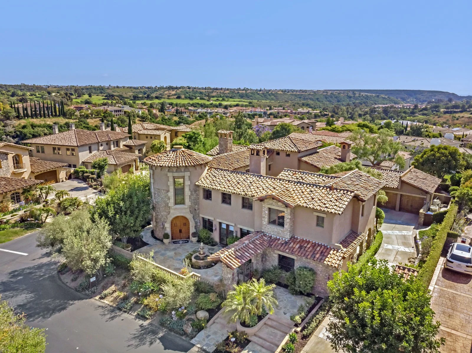 Aerial view of a luxurious neighborhood with large, Mediterranean-style homes featuring tiled roofs, lush greenery, and a winding road in the foreground under a clear blue sky.