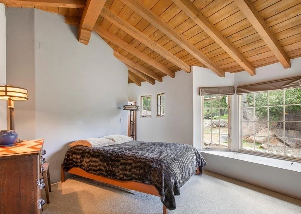 Bedroom with a wooden vaulted ceiling, bed with dark blanket, window seat with large windows, and a nightstand with a lamp.