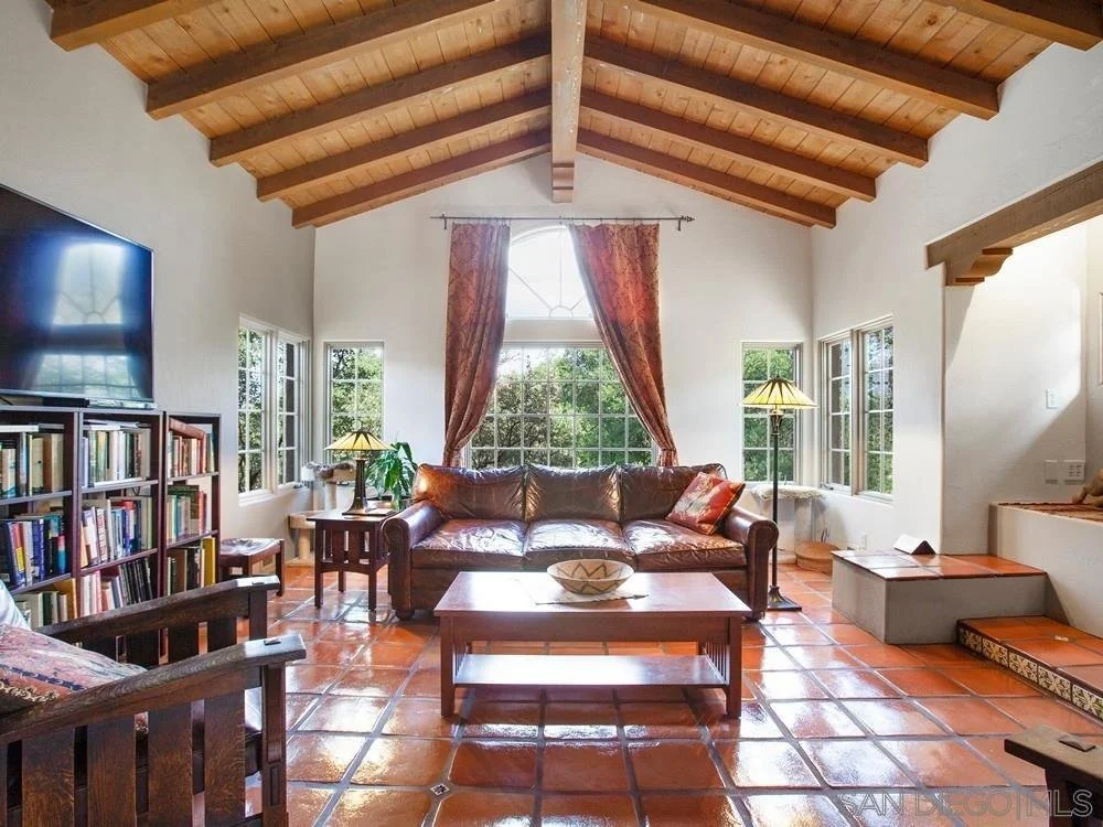Living room with wooden vaulted ceiling, beige walls, terracotta tile floor, leather sofa, wooden coffee table, bookshelf, large windows with curtains, and area lamps.