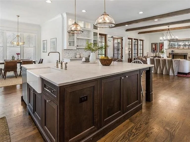 Open-concept kitchen and dining area with hardwood floors, a large island with dark wood cabinets, pendant lighting, a white marble countertop, and a living room with a fireplace and glass doors in the background.