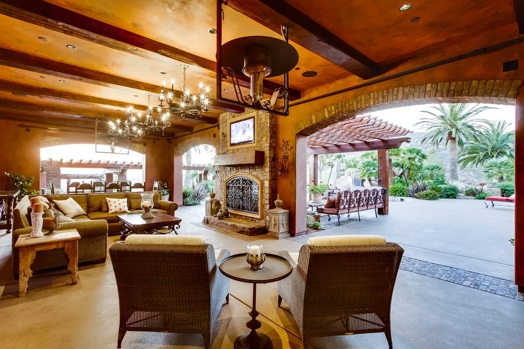 Interior of a cozy lounge area with a fireplace, wicker chairs, sofas, a chandelier, and an outdoor patio with palm trees.