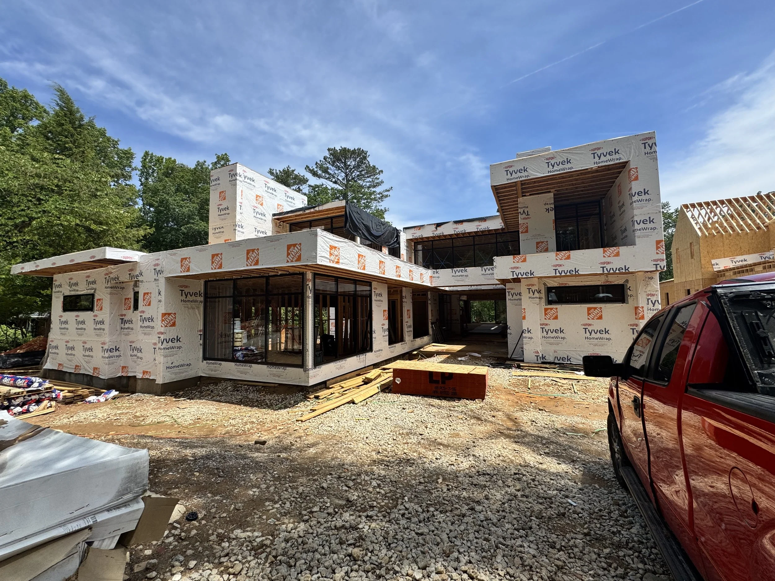 A house under construction with wooden framing and exterior insulation wrapped in Tyvek HomeWrap. There are trees and a blue sky in the background, and construction materials and debris on the ground.