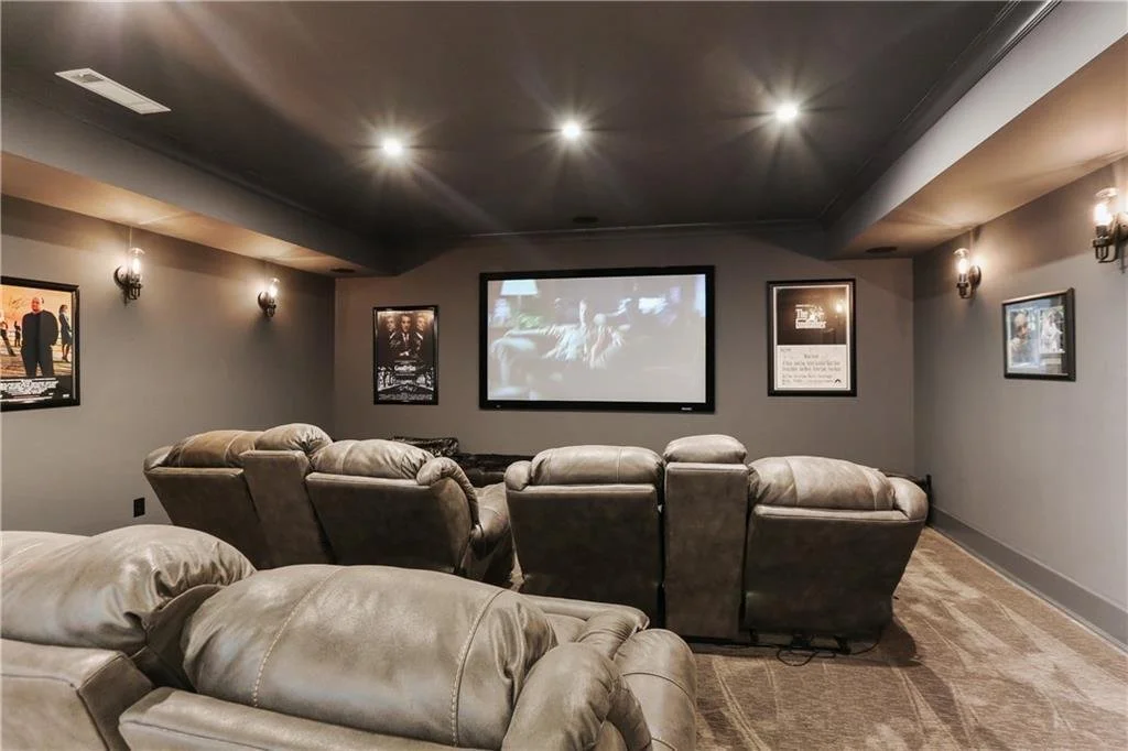 A cozy home theater room with several reclining chairs facing a large screen, framed movie posters on the walls, and soft lighting.