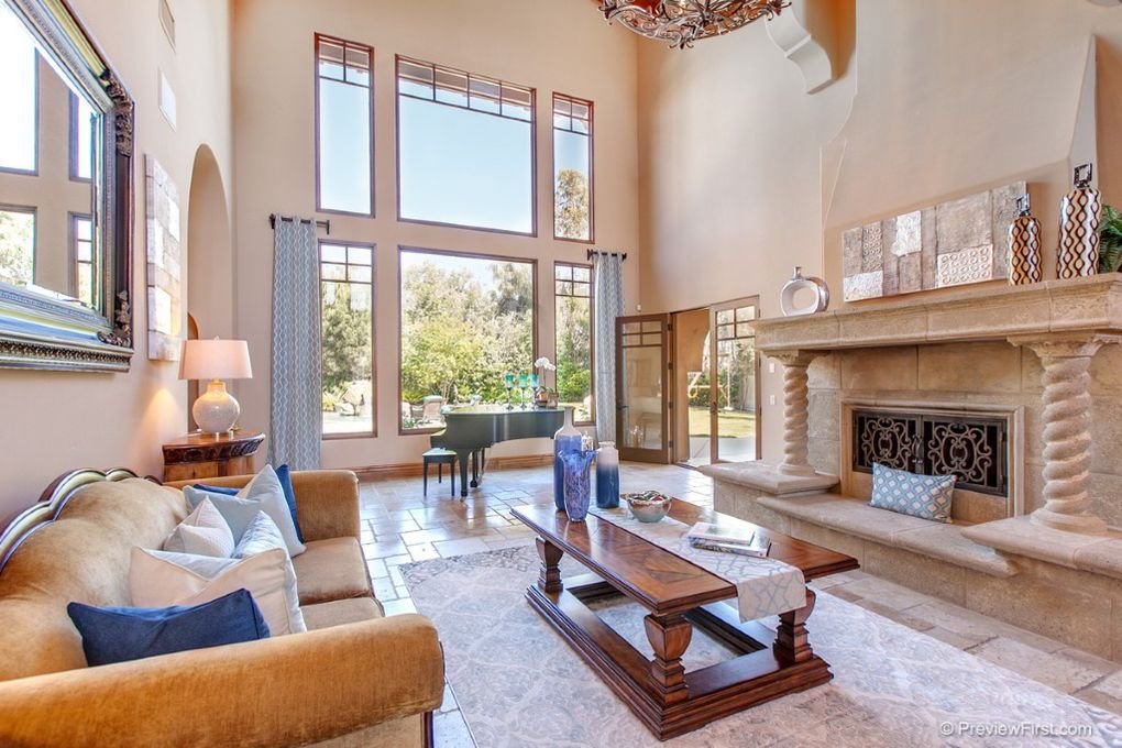 Living room with large floor-to-ceiling windows, a beige sofa with white and blue pillows, a wooden coffee table with decorative blue vases, a stone fireplace with vases and a small pillow, a piano by the window, and a large mirror on the wall.