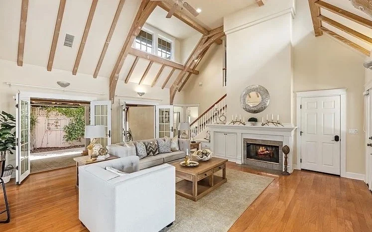 Living room with wooden vaulted ceiling, a fireplace, white walls, and hardwood floors. Features a white sofa, a wooden coffee table, table lamps, and glass doors leading outside.