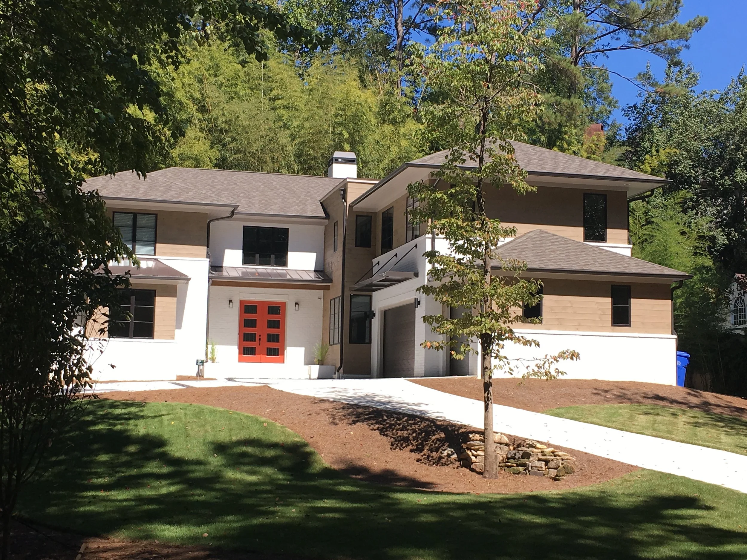 Modern house with beige and white exterior, black window frames, a red front door, surrounded by lush trees and a manicured lawn under a clear blue sky.
