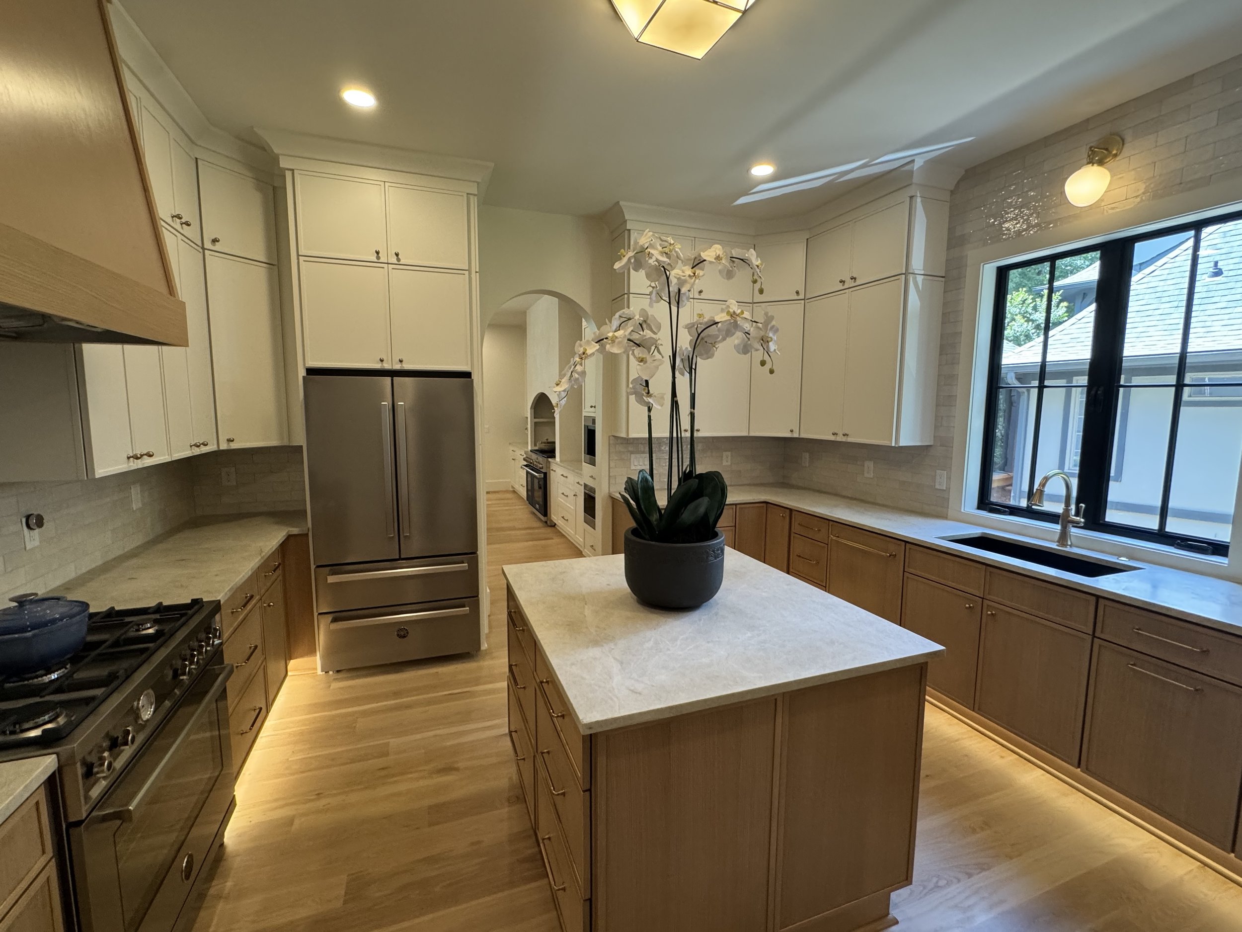 A spacious kitchen with white upper cabinets, light wood lower cabinets, and a white marble countertop island. There is a stainless steel refrigerator, a gas stove, and large windows with black frames allowing natural light. A potted orchid sits on t