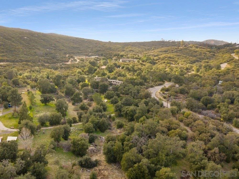 An aerial view of a hilly landscape with winding roads, dense trees, and scattered houses under a partly cloudy sky.